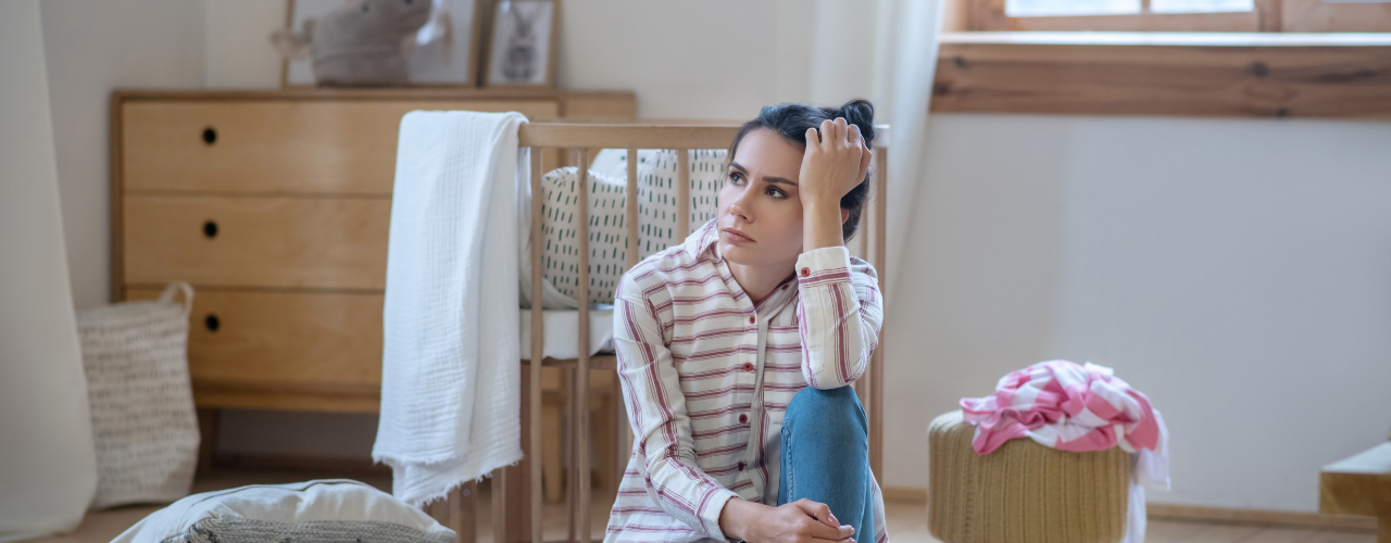 Mulher sentada no chão, com expressão cansada e ansiosa, apoiando a cabeça na mão, num quarto simples — representando exaustão, stress e o momento em que o corpo pede pausa.