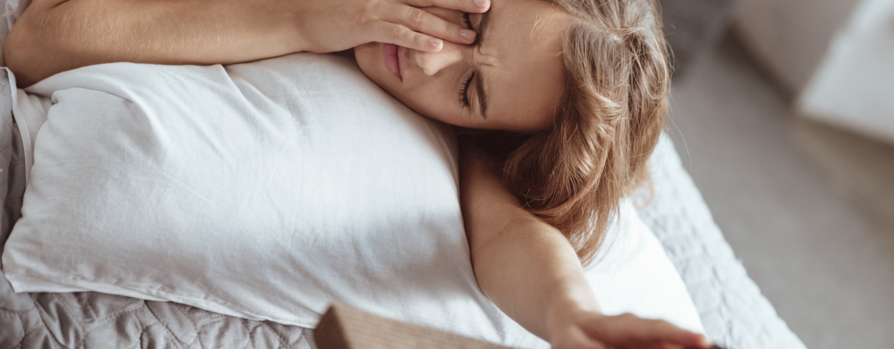 Mulher deitada na cama de manhã, mão a pressionar os olhos, a desligar o despertador com uma expressão de cansaço apesar de ter dormido a noite toda